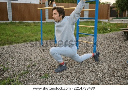 Handsome middle-aged dark-haired Caucasian sporty man in active wear, exercising outdoor with suspensions straps, doing lunges on the sportsground. Bodyweight training. Sport and healthy lifestyle