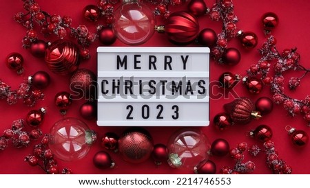 Marry Christmas banner concept. White board with the text Marry Christmas 2023 among Christmas red decor, balls and berries on a red background, top view, flat lay, copy space
