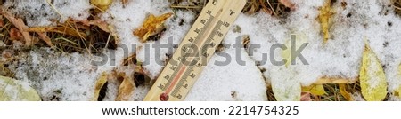 Thermometer with a Celsius and Fahrenheit scale lies among the yellow autumn foliage and freshly fallen snow. Temperature is below zero. Cold snap, frosts and the first snow. Web banner. Close-up Royalty-Free Stock Photo #2214754325