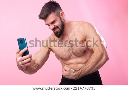 Strong and muscled bodybuilder taking a selfie with his smartphone of his shirtless torso, showing off his gains to friends on social media