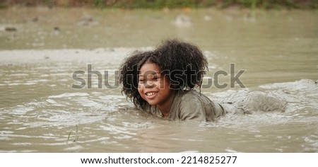 Happy african-american children girl playing in wet mud puddle during raining in rainy season.