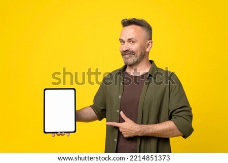 smiling handsome bristle with mustach mature man in green shirt pointing with finger and thumb up to his tablet pc with white mock-up screen holding in hand over yellow background