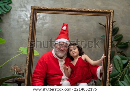 Portrait of happy Santa Claus wearing red costume and cute little girl in red dress sitting on Santa lap in wooden picture frame