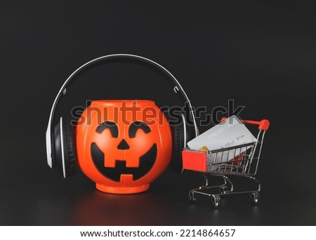 Front view of  plastic Halloween pumpkin  bucket covere with headphones  isolated  on black  background  with shopping cart or trolley and credit card. Halloween holiday shopping  concept.