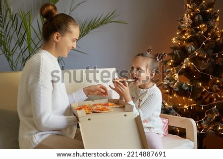 Horizontal shot of happy positive family, mother and daughter sitting on sofa in decorated festive living room with Xmas tree, eating junk food, enjoying tasty pizza on holiday.