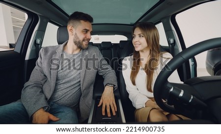 Loving husband is giving car key to his happy wife while sitting inside new automobile with nice interior and leather seats. Excited woman is kissing him holding steering wheel.