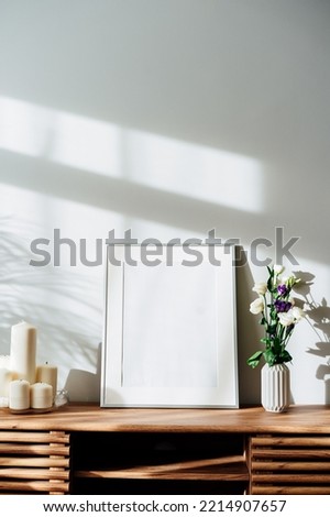 Modern minimalist Scandinavian style interior with white poster mockup, candles and flowers in vase on a wooden console under sunlight and home plants shadows on a white gray wall. Selective focus