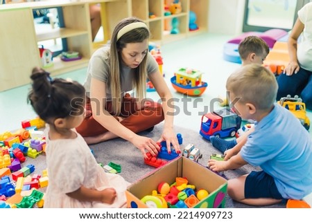 young blond teacher building with blocks for her beloved kids in the nursery, sitting on the floor numerous toys in the background. High quality photo