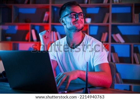 Positive emotions. Man in glasses and white shirt is sitting by the laptop in dark room with neon lighting.