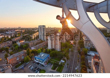 Urban climbing: free solo rock climber hanging on element of roof of high building at sunset. Panoramic view of city at background Royalty-Free Stock Photo #2214947623
