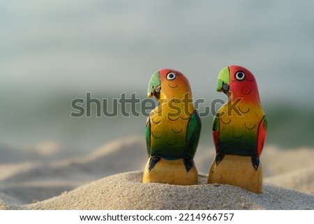 Couple of toy parrots. They have red and yellow feathers on face, chest, crown, green on body. Birds caress each other like wife and husband. Apparently these are lovebirds. They are sitting  on beach
