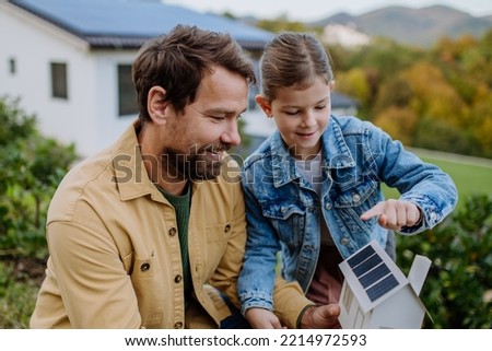 Little girl with her dad holding paper model of house with solar panels, explaining how it works.Alternative energy, saving resources and sustainable lifestyle concept. Royalty-Free Stock Photo #2214972593