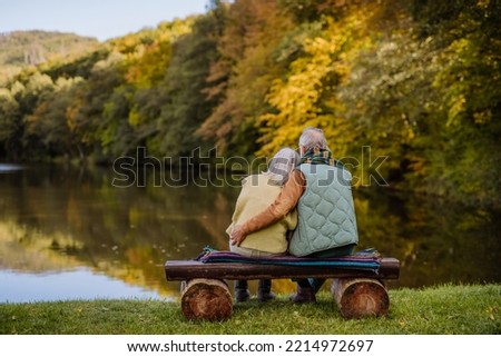 Senior couple in love sitting together on bench looking at lake, during autumn day. Rear view. Royalty-Free Stock Photo #2214972697