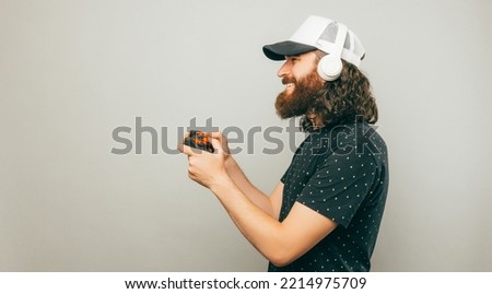 Smiling bearded curly man is playing with a joystick while wearing headphones. Banner studio shot over light grey background.