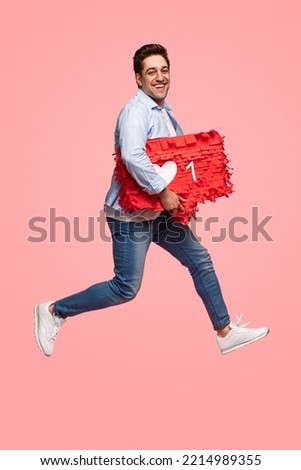 Side view of cheerful young ethnic male with dark hair in casual clothes, smiling while running with red box with heart symbol against pink background