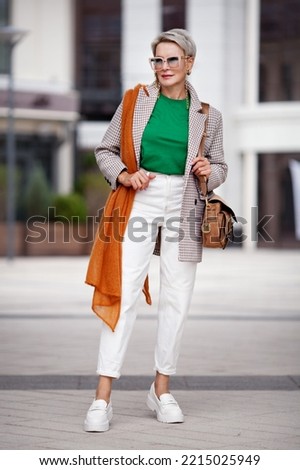 Fashion model 50 years stands street city full length, wears stylish colorful outfit, color T-shirt, jacket, bag, white pants and loafers, knitted scarf. Street style Royalty-Free Stock Photo #2215025949