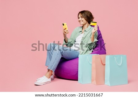 Full body young woman wear green shirt sit in bag chair at shopping paper bags hold credit bank card mobile phone order delivery isolated on plain pastel pink background. Black Friday sale buy concept Royalty-Free Stock Photo #2215031667