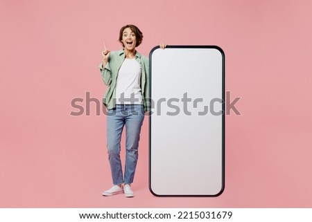 Full body young proactive woman wear green shirt white t-shirt near big huge blank screen mobile cell phone smartphone with mockup area point finger up isolated on plain pastel light pink background