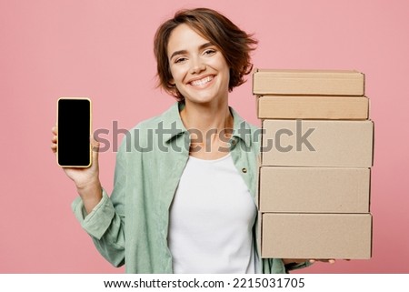 Young woman wear green shirt white t-shirt hold in hand use mobile cell phone with black screen area, brown clear blank craft paper cardboard box mock up isolated on plain pastel light pink background