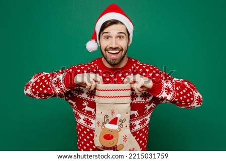 Merry exultant young man 20s wear red warm knitted sweater Santa hat posing hold in hand show stocking sock isolated on plain dark green background studio. New Year 2023 holiday celebration concept