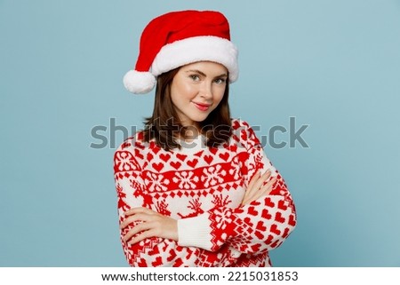Merry fun smiling cool woman 20s wearing warm red knitted sweater Santa hat posing hold hands crossed folded isolated on plain pastel light blue cyan background. Happy New Year 2023 holiday concept