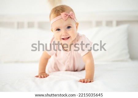 a little cute healthy girl up to a year old in a pink bodysuit made of natural fabric is sitting on a bed on white bed linen in the bedroom, looking at the camera, the baby is at home Royalty-Free Stock Photo #2215035645