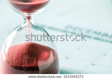 Close up of red sand slipping away through hourglass, time running out, December calendar out of focus in background, end of year, no more time Royalty-Free Stock Photo #2215042275