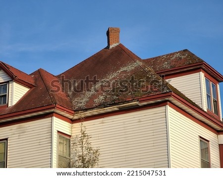 A red shingled rooftop that has mold and mildew covering most of the weathered shingles with lots of missing pieces on the shingles. Royalty-Free Stock Photo #2215047531