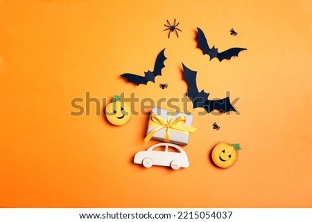 Wooden toy car with gift on the roof, bats and pumpkins on orange background. Flat lay Halloween background with copy space.