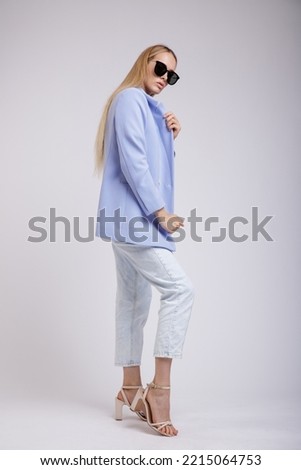 High fashion photo of a beautiful elegant young woman in a pretty blue jacket, blazer, denim jeans, stylish sunglasses posing over white, soft gray background. Studio Shot. Slim Figure, Blonde