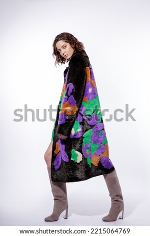 High fashion photo of a beautiful elegant young woman in a pretty black fur coat with colorful patterns, suede boots posing over white background. Make up, hairstyle. Slim figure.
