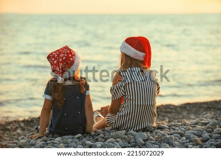 two sister girls in santa claus hats sit and chat merrily on the seashore at sunset