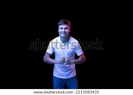 Portrait of young man in white T-shirt posing isolated over black background in neon light. Looking happy and cheerful. Concept of emotions, facial expression, lifestyle, fashion, youth