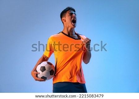 Portrait of young expressive man, football player posing isolated over blue studio background in neon light. Winning game. Concept of sport, team game, action, motion. Copy space for ad, poster Royalty-Free Stock Photo #2215083479