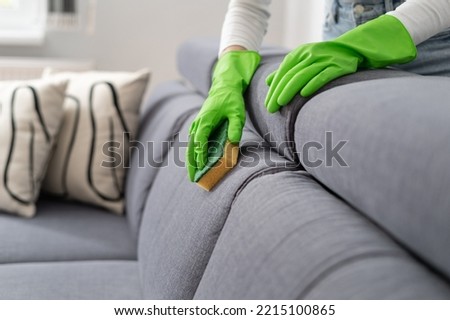 cropped shot of female hands in green rubber gloves wash couch upholstery with sponge in living room at modern apartment, housework concept Royalty-Free Stock Photo #2215100865