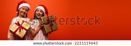 cheerful preteen girls in santa hats holding christmas gifts isolated on orange, banner Royalty-Free Stock Photo #2215119643