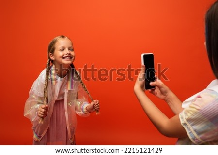 funny girl holding her pigtails near friend taking photo on smartphone isolated on orange