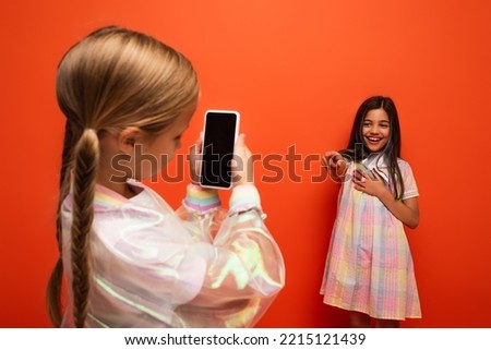 girl with pigtails taking photo of laughing friend pointing with finger isolated on orange