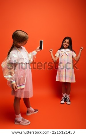 full length of happy girl showing win gesture near friend taking photo on mobile phone on orange background