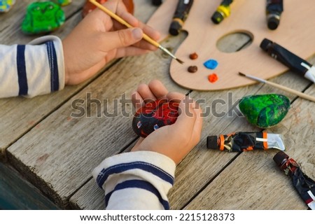 A detailed picture of a child's hands drawing a ladybug on a stone with acrylic paints. Home hobbies are authentic. Artwork on stones.