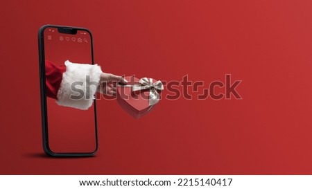 Santa Claus hand holding a heart shaped Christmas gift and coming out from a smartphone screen, blank copy space Royalty-Free Stock Photo #2215140417