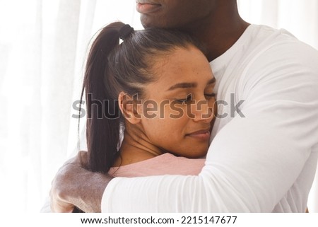 Happy diverse couple embracing by window at home, smiling. Happiness, domestic life, love and inclusivity concept.