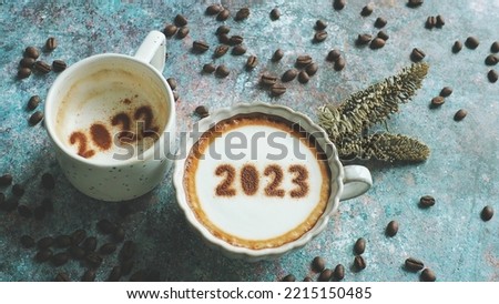 Goodbye 2022, Hello 2023 theme coffee cups with number 2023 and 2022 over frothy surface on rustic blue background with coffee beans and dried pine branches. Holidays food art for Happy New Year.