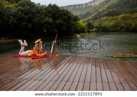 The girl artist paints a painting with a picturesque view of the lake in the rain. The woman in wearing bright yellow clothes
