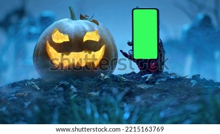 Zombie hand holds smartphone with green screen out of grave near Glowing Halloween pumpkin in the cemetery. Holiday event halloween concept.
