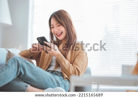 Happy young asian woman browsing surfing wireless internet on mobile phone while sitting a couch in living room at home, Shopping online via website Royalty-Free Stock Photo #2215165689