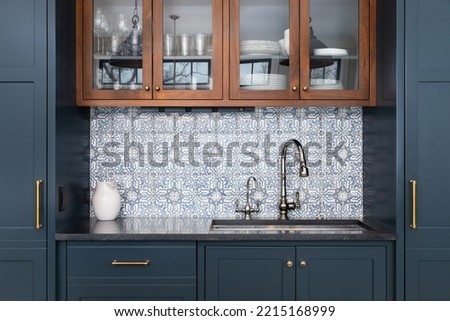 A kitchen sink with a beautiful pattern tiled backsplash with a chrome faucet, black granite countertops, and surrounded by blue and wood cabinets.