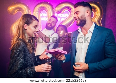 Beautiful couple celebrating New Year or birthday at night club and having fun together while exchanging gifts. Diverse friends with sparklers partying in the background. Love, romance and presents.