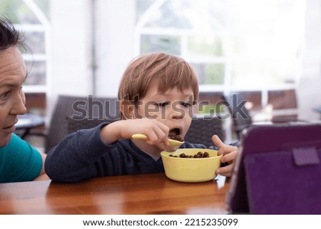 Little boy sit on chair at table and with interest watch cartoon on tablet computer with mother. Kid of kindergarten age open mouth to eat chocolate balls with milk. Ready breakfast, gadget addiction.