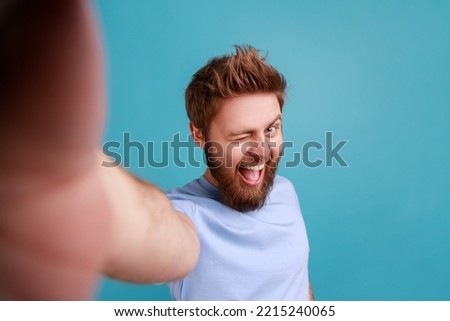 Portrait of excited optimistic bearded man taking selfie POV, winking to camera, being in excellent mood, flirting, point of view photo. Indoor studio shot isolated on blue background.